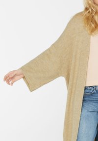 Beige gestrickte Strickjacke mit weiten Ärmeln, offenem Vorderdesign und weicher Textur, die über einem hellen Oberteil und hellblauen Jeans getragen wird.
