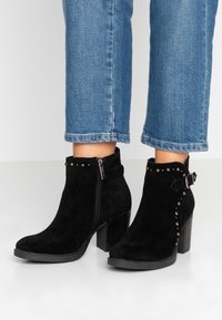 Botas de tobillo de ante negro con tacones en bloque, cremallera lateral, tachuelas decorativas y hebilla, combinadas con jeans azul cortos.