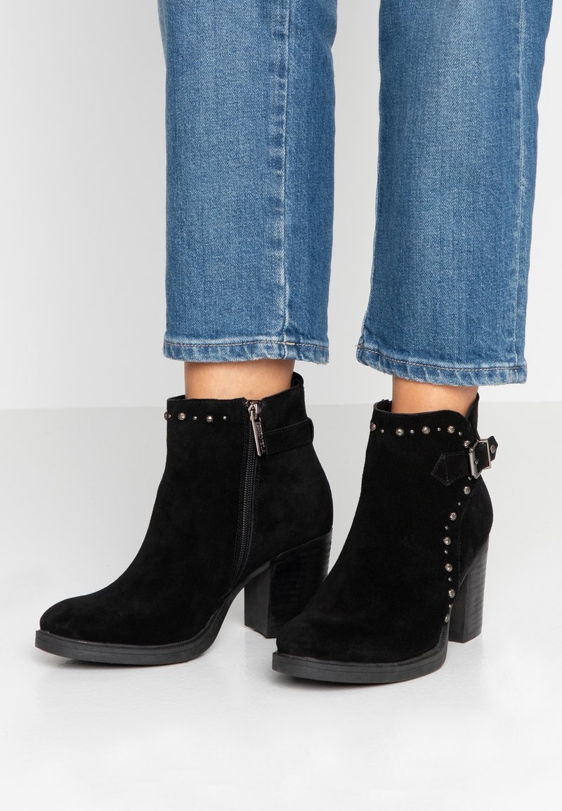 Botas de tobillo de ante negro con tacones en bloque, cremallera lateral, tachuelas decorativas y hebilla, combinadas con jeans azul cortos.