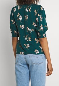 Frau trägt eine türkisfarbene Bluse mit braunem und weißem Blumen- und Schmetterlingsmuster sowie eine hellblaue Jeans, von hinten betrachtet.