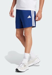 Blauwe sportshorts met drie witte verticale strepen aan elke kant. Draagt witte sokken en grijs-witte sportschoenen.