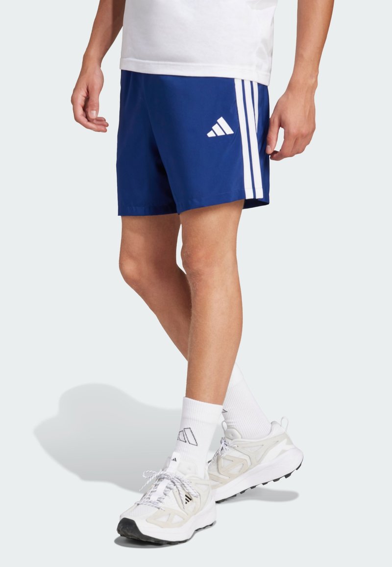 Blauwe sportshorts met drie witte verticale strepen aan elke kant. Draagt witte sokken en grijs-witte sportschoenen.