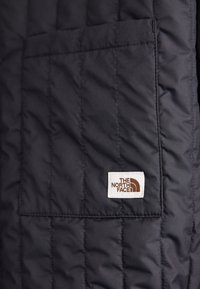 Quiltet mørkeblå jakke med en frontlomme og et hvidt mærke, der viser "The North Face"-logoet i brunt. Glat, syntetisk tekstur.