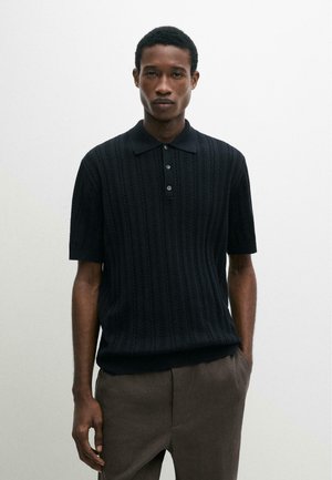 Jeune homme aux cheveux courts portant un polo noir à manches courtes texturé et un pantalon marron, debout devant un fond blanc uni.