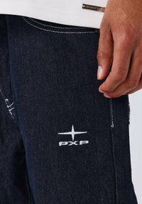 Zwarte denim jeans met witte stiksels en een geborduurd logo met de tekst "PXP" op de linkerdij. Bevat een close-up van een hand die de stof vasthoudt.