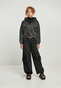 Sudadera recortada de patrón de mármol negro con cremallera delantera y capucha, combinada con pantalones cargo negros que presentan bolsillos laterales y un acabado texturizado.