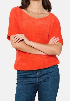 Kobieta w jasnopomarańczowym, puszystym swetrze z krótkim rękawem i niebieskich dżinsach, z założonymi skrzyżowanymi rękami na torsie, na jednolitym tle.