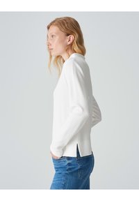 Weißes Langarmoberteil mit Waffelstruktur, Rundhalsausschnitt, seitlichen Schlitzen, kombiniert mit blauen Jeans. Einfaches und enganliegendes Design.