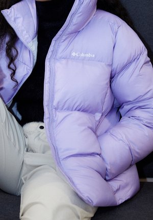 Henkilö pukeutunut vaaleanpunaiseen Columbia-toppatakkiin mustan paidan ja vaaleanbeigen housujen päällä, istuu käsi koukussa.