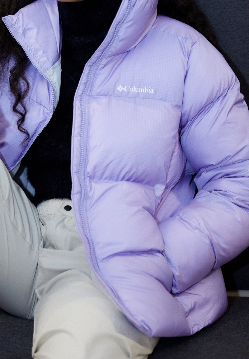 Personne portant une doudoune Columbia violet clair par-dessus un haut noir et un pantalon beige clair, assise avec un bras plié.