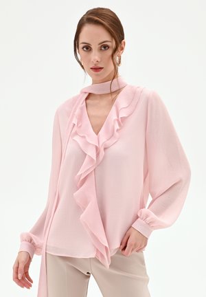 Donna che indossa una camicetta rosa chiaro con maniche lunghe, scollo a V arricciato e una sciarpa da collo abbinata annodata, abbinata a pantaloni beige.