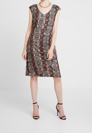 Woman wearing sleeveless knee-length brown snake print dress with black ankle-strap high heels and gold chain necklace, standing on plain background.