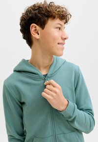Turquoise capuchonvest met een rits aan de voorkant, kangoeroezakken en geribbelde boorden. Zachte stof en capuchon met een gladde textuur.