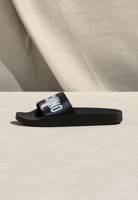 Μαύρο σανδάλι με πλατιά λουρί που φέρει λευκά γράμματα που σχηματίζουν τη λέξη «MOSCHINO», τοποθετημένο σε μπεζ επιφάνεια με υφασμάτινο φόντο.