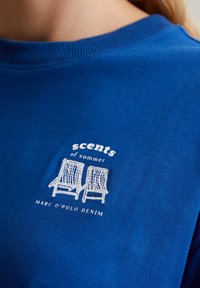 Blaues Baumwoll-T-Shirt mit einer weißen gestickten Grafik von zwei Stühlen, darunter der Text "Düfte des Sommers" und "MARC O'POLO DENIM".