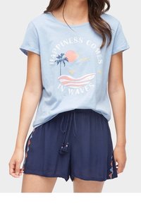 Kvinna som bär en ljusblå T-shirt med palmer, sol, fåglar och texten "Lycka kommer i vågor", ihop med marinblå shorts med snöre och sidobroderi.