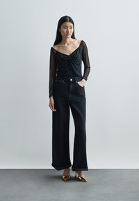 Black sheer long-sleeve top with a draped neckline, paired with high-waisted wide-leg black jeans and gold pointed-toe heels.