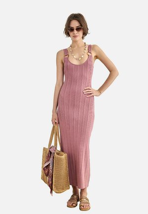 Mujer con un vestido largo rosa sin mangas y acanalado con detalles de anillos, gafas de sol, collar, sandalias y sosteniendo un bolso tejido con una bufanda.