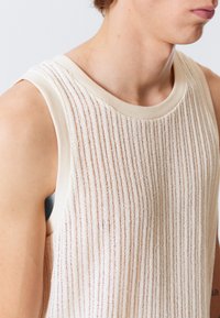 Strikket ermeløs topp i kremfarge med vertikale striper. Har en rund halslinning og litt bredere skuldersnitt. Myk og pustende stoff.