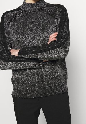 Person with crossed arms wearing a black and silver glittery knit sweater with textured black stripes on the sleeves and black pants.