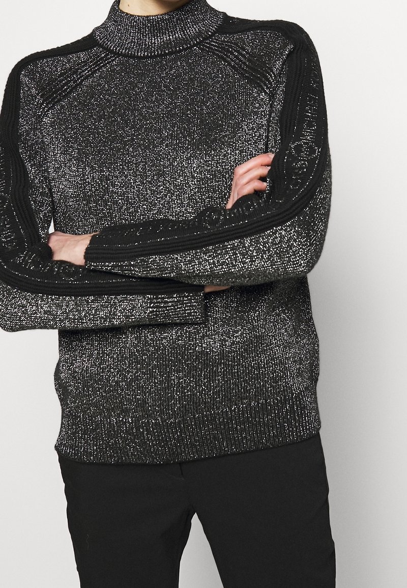Personne les bras croisés portant un pull en maille noir et argenté pailleté avec des rayures noires texturées sur les manches et un pantalon noir.