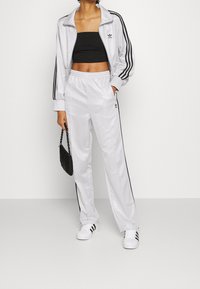 White tracksuit featuring a cropped jacket and relaxed-fit pants with black stripes. Textured fabric and logo detail on the chest and pant.