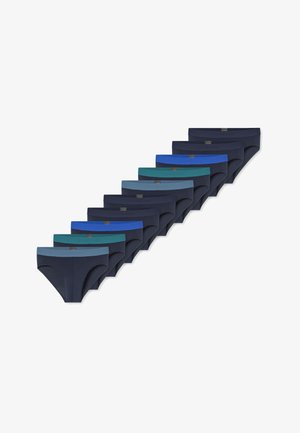 Fit jonge man met kort haar, gekleed in marineblauwe slips met een grijze tailleband, poseert met zijn handen achter zijn hoofd tegen een effen achtergrond.