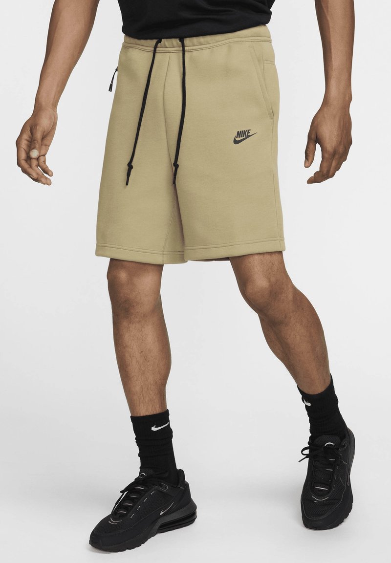 Béžové Nike šortky s elastickým páskem a šňůrkami na stažení; černé logo na levé straně; nošeno s černými ponožkami a teniskami.
