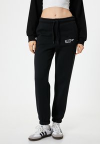 Schwarze Jogginghose aus Baumwollmischung, mit einem Kordelzugbund, gerippten Säumen und weißem Drucktext an der Hüfte. Kombiniert mit weißen Sneakers.