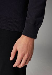 Tumesinine kudumipusa, mille mansetid on soonilised, näidatud koos käega. Tekstuur on sile ning materjal näib pehme ja veniv.