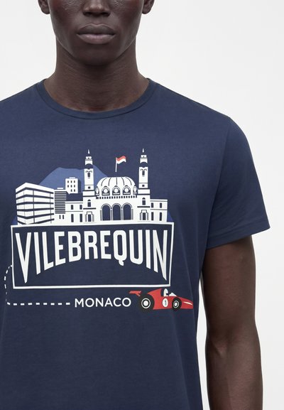 Άνδρας που φοράει μπλε σκούρο t-shirt με λευκό και κόκκινο σχέδιο που δείχνει κτίρια του Μονακό, ένα κόκκινο αγωνιστικό αυτοκίνητο και το κείμενο "Vilebrequin Monaco."