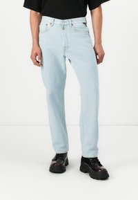 Helle blaue Jeans aus Denim mit geradem Schnitt, fünf Taschen, Knopfverschluss und subtilen Nahtdetails; getragen mit schwarzen Schnürstiefeln.