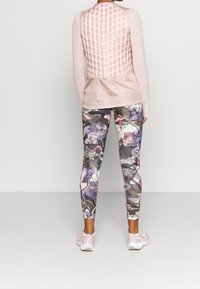 Kobieta w różowej pikowanej kamizelce, długim rękawie z fakturowanym wzorem, legginsach w kwiatowy nadruk oraz różowych butach do biegania, stojąca na tle jednolitego tła.