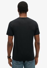 Zwarte katoenen t-shirt met korte mouwen, ronde halslijn en een gladde textuur, van achteren weergegeven. Jeans zichtbaar bij de taille.