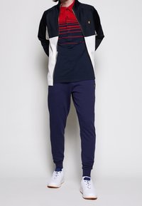 Giacca a zip blu navy e bianca sopra una polo a righe rosse e blu navy, abbinata a pantaloni jogger blu navy e sneaker bianche con suole marroni.