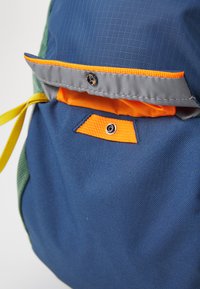 Mochila multicolor con un tejido azul texturizado, un bolsillo interior naranja, ribetes grises y un cordón amarillo de cierre.