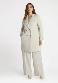 Beige double-breasted coat with button accents, tailored fit, and two side pockets, paired with matching wide-leg trousers and light shoes.