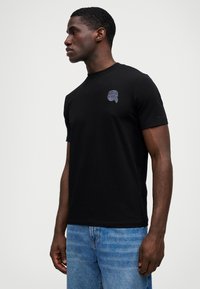 Μαύρη βαμβακερή T-shirt με κοντό μανίκι και στρογγυλή λαιμόκοψη. Περιλαμβάνει μια μικρή γραφική απεικόνιση στο αριστερό στήθος που απεικονίζει ένα πρόσωπο.