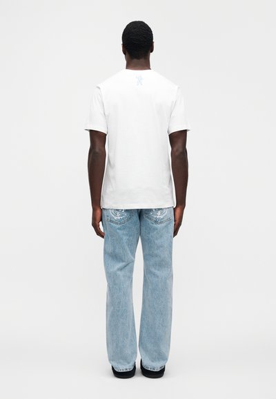 Άσπρο βαμβακερό μπ λουζάκι με κοντά μανίκια, με μπλε γραφικό στο πάνω μέρος της πλάτης. Συνδυάζεται με ανοιχτό μπλε τζιν παντελόνι με χαλαρή εφαρμογή.