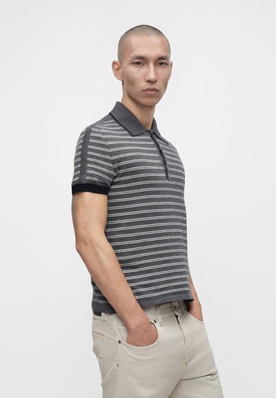 Homme aux cheveux courts portant un polo rayé gris et noir et un pantalon beige clair, les mains dans les poches, debout devant un fond uni.