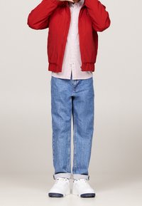 Niño que lleva una chaqueta roja, una camisa de botones a rayas, vaqueros azules con puños y zapatillas blancas, de pie con las manos cerca de la cara.