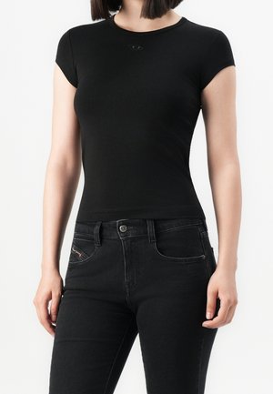 Femme portant un t-shirt noir ajusté à manches courtes et un jean noir, visible du cou aux hanches sur un fond blanc uni.