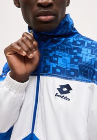 Χέρι άνδρα σηκώνοντας το φερμουάρ σε μπλε και λευκό μπουφάν Lotto με γεωμετρικά σχέδια και λογότυπο στο στήθος.