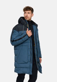 Blau und schwarz gepolsterter langer Mantel mit Kapuze, der über einen Reißverschluss, seitliche Taschen und einen Kordelzug in der Kapuze für Anpassungsmöglichkeiten verfügt.