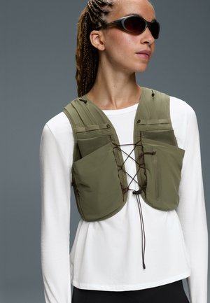 Femme aux cheveux tressés portant des lunettes de soleil noires, une chemise blanche à manches longues et un gilet utilitaire vert olive avec des poches avant et une fermeture à lacets.