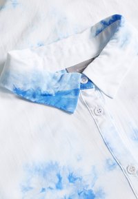Camicia bianca con motivo tie-dye azzurro chiaro sul colletto, dotata di una chiusura con bottoni e due bottoni bianchi vicino al collo.