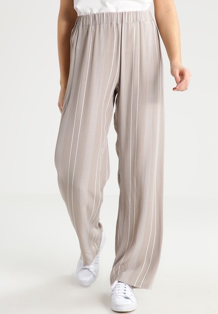 Pantalons larges beige clair avec des rayures verticales blanches. Présente une taille élastique et une texture lisse, associés à des baskets blanches.