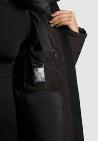 Schwarze Steppjacke mit sichtbarem Reißverschlussfach, Textillabel und kontrastierenden, dunkelbraunen Seitenteilen. Glatte und matte Oberflächen.