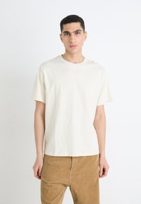 T-shirt en coton couleur crème à manches courtes et à col rond. Présente un logo discret sur la poitrine gauche. Porté avec un pantalon en velours côtelé marron clair.