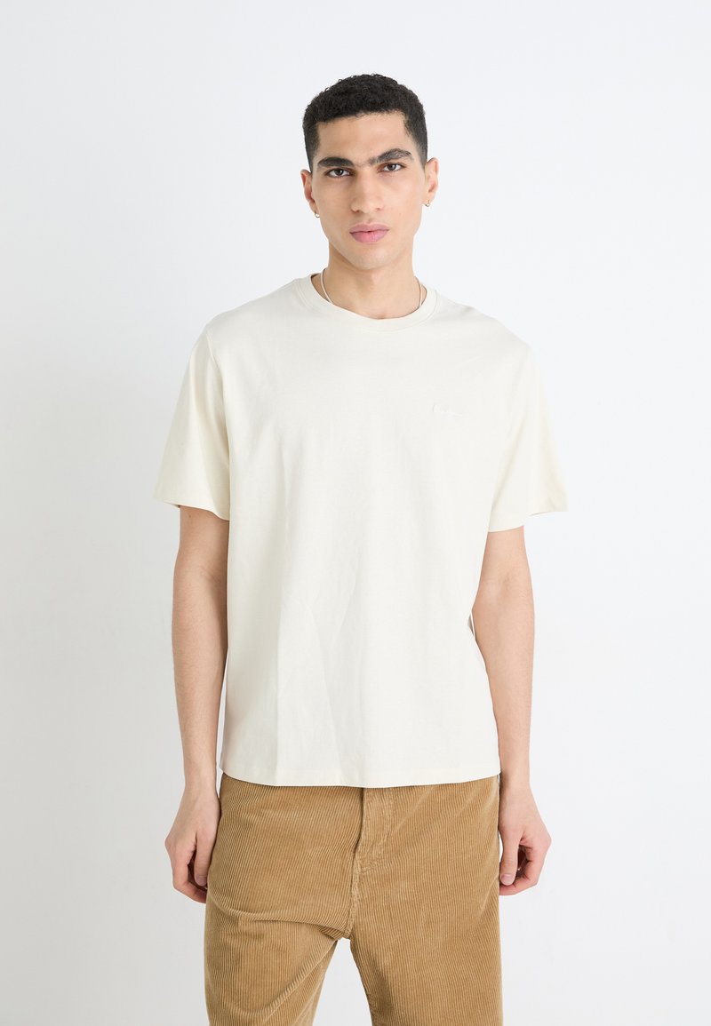 T-shirt en coton couleur crème à manches courtes et à col rond. Présente un logo discret sur la poitrine gauche. Porté avec un pantalon en velours côtelé marron clair.